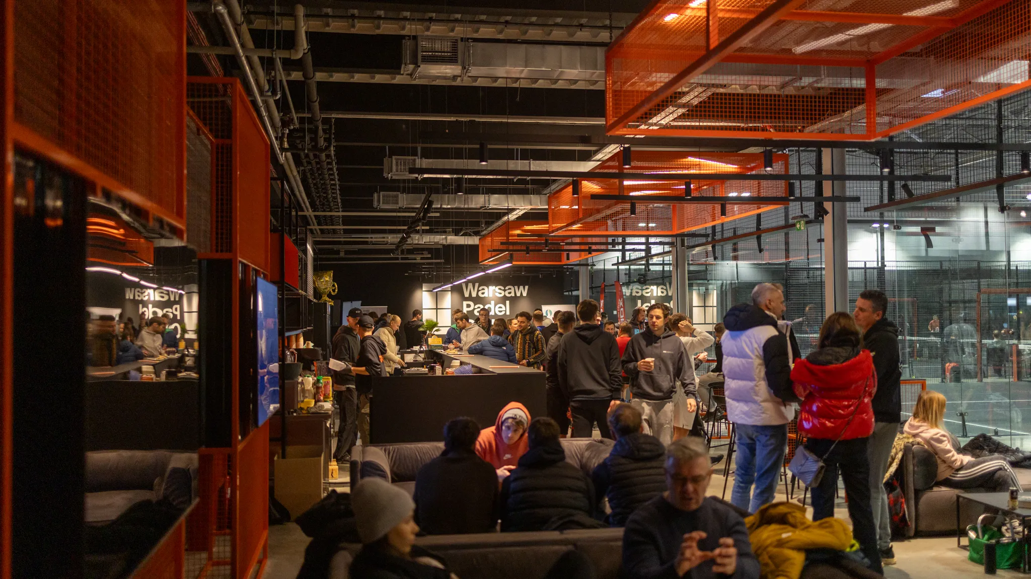 Indoor padel club with people socializing near a bar area and sitting on couches, with padel courts visible in the background under orange ceiling fixtures.