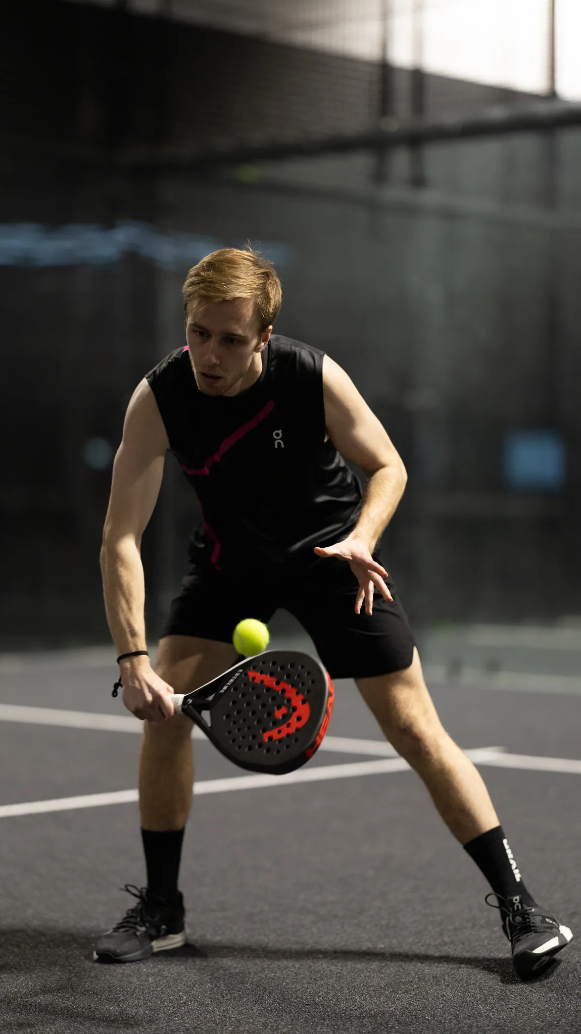 Young man in black sportswear playing paddle tennis, hitting a yellow ball with a paddle racket on an indoor court.
