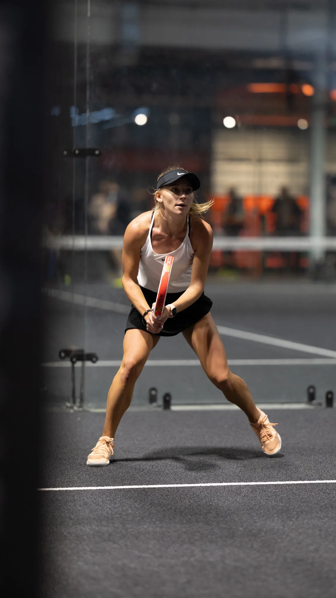 Female padel player in a white tank top and black skirt preparing to hit a ball on an indoor court.