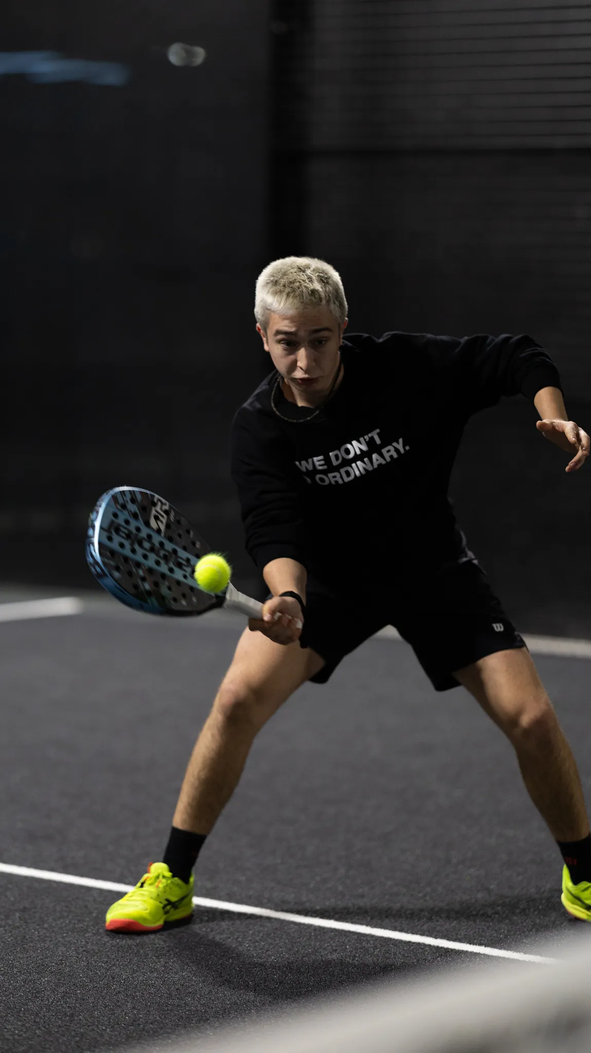 Padel player in black outfit and neon yellow shoes hitting a ball with a blue paddle on a dark court.