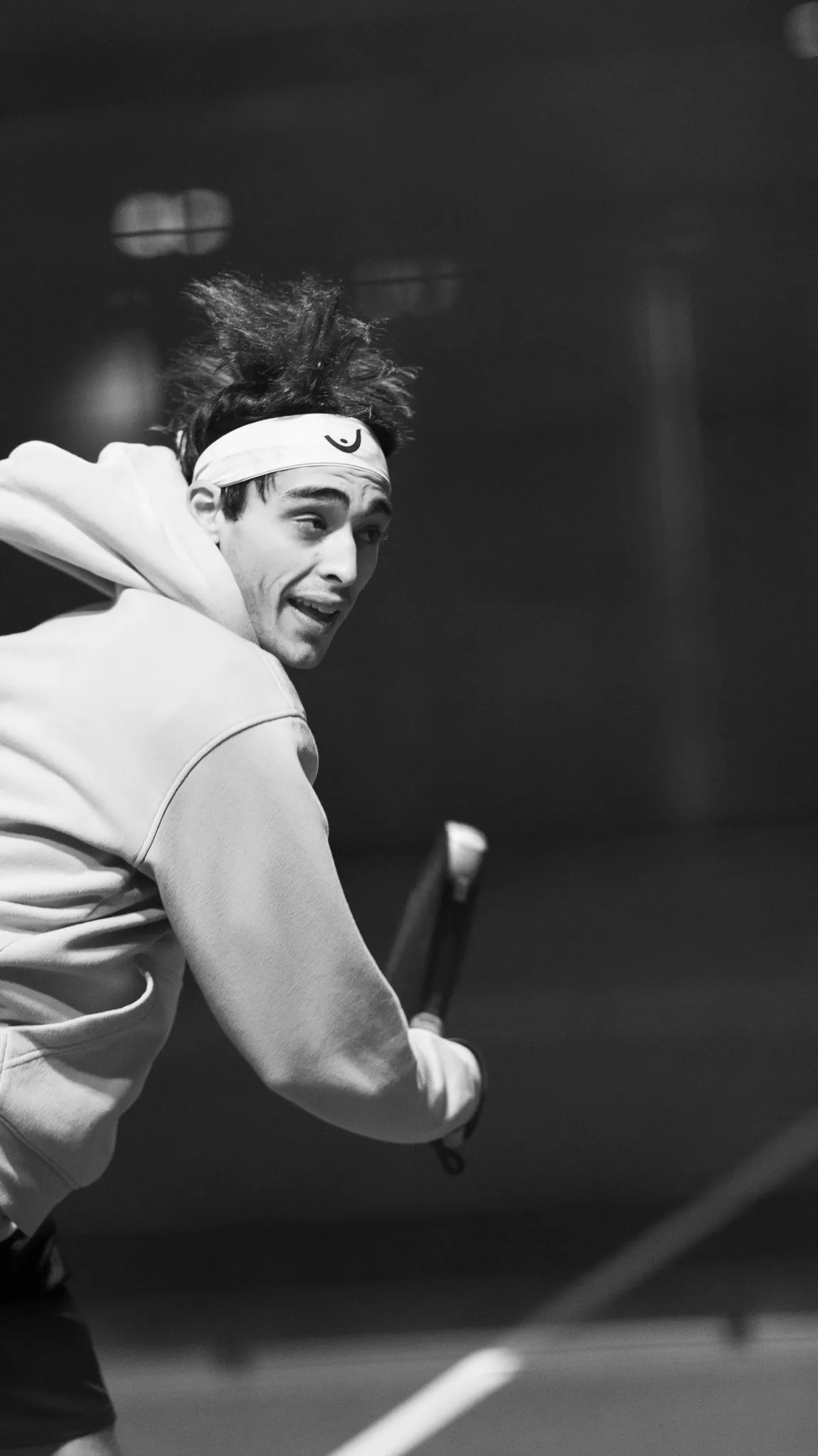 Male padel player in a headband and hoodie holding a racket, preparing to hit a ball on an indoor court.