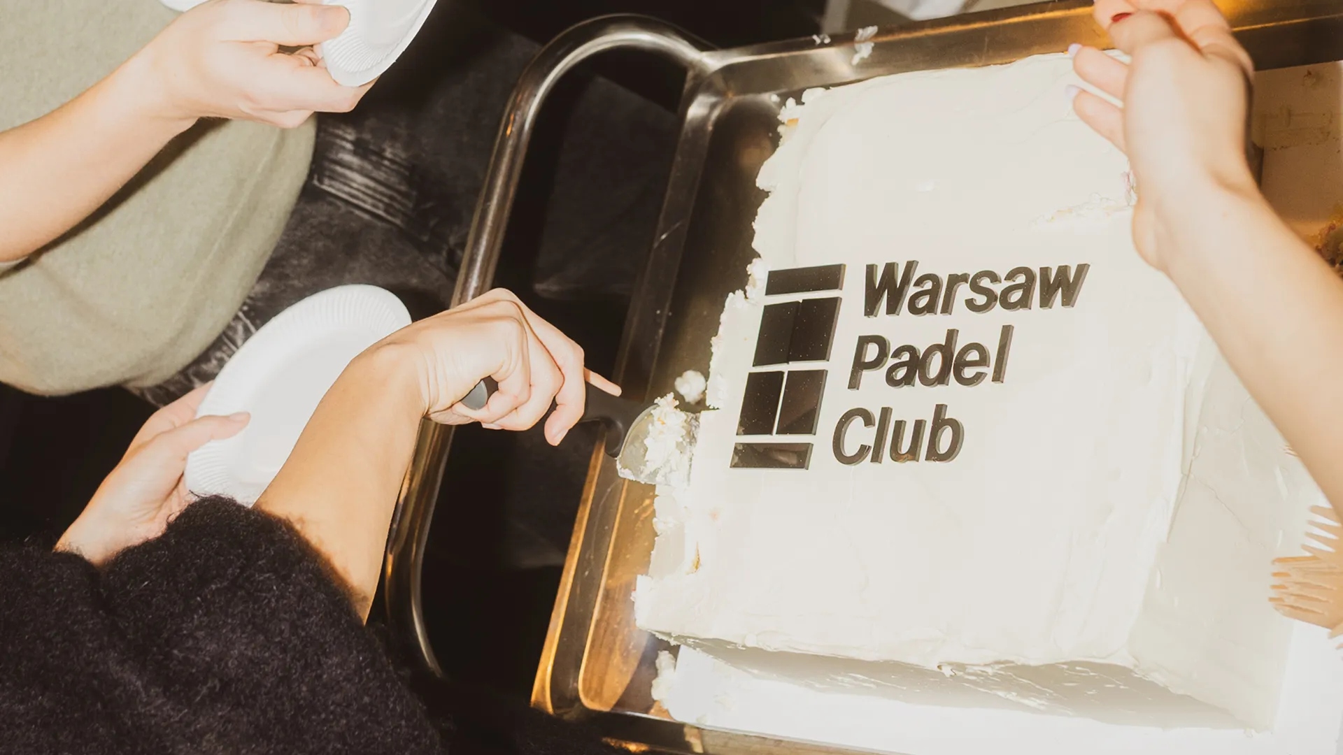 Hands cutting and serving a white cake with Warsaw Padel Club logo on top.