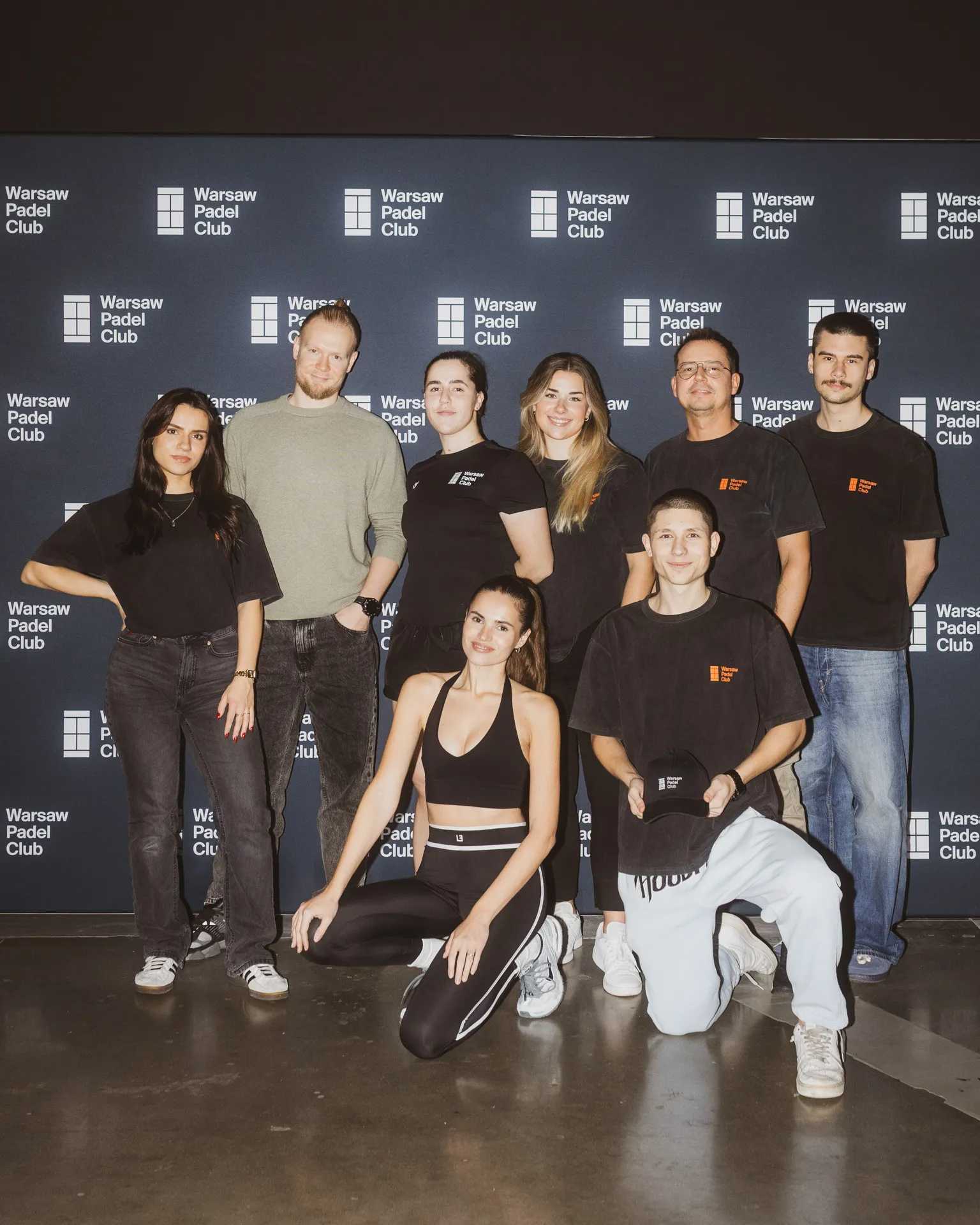 Group of eight people posing in front of a Warsaw Padel Club backdrop, with some standing and some kneeling, dressed in casual and sportswear.