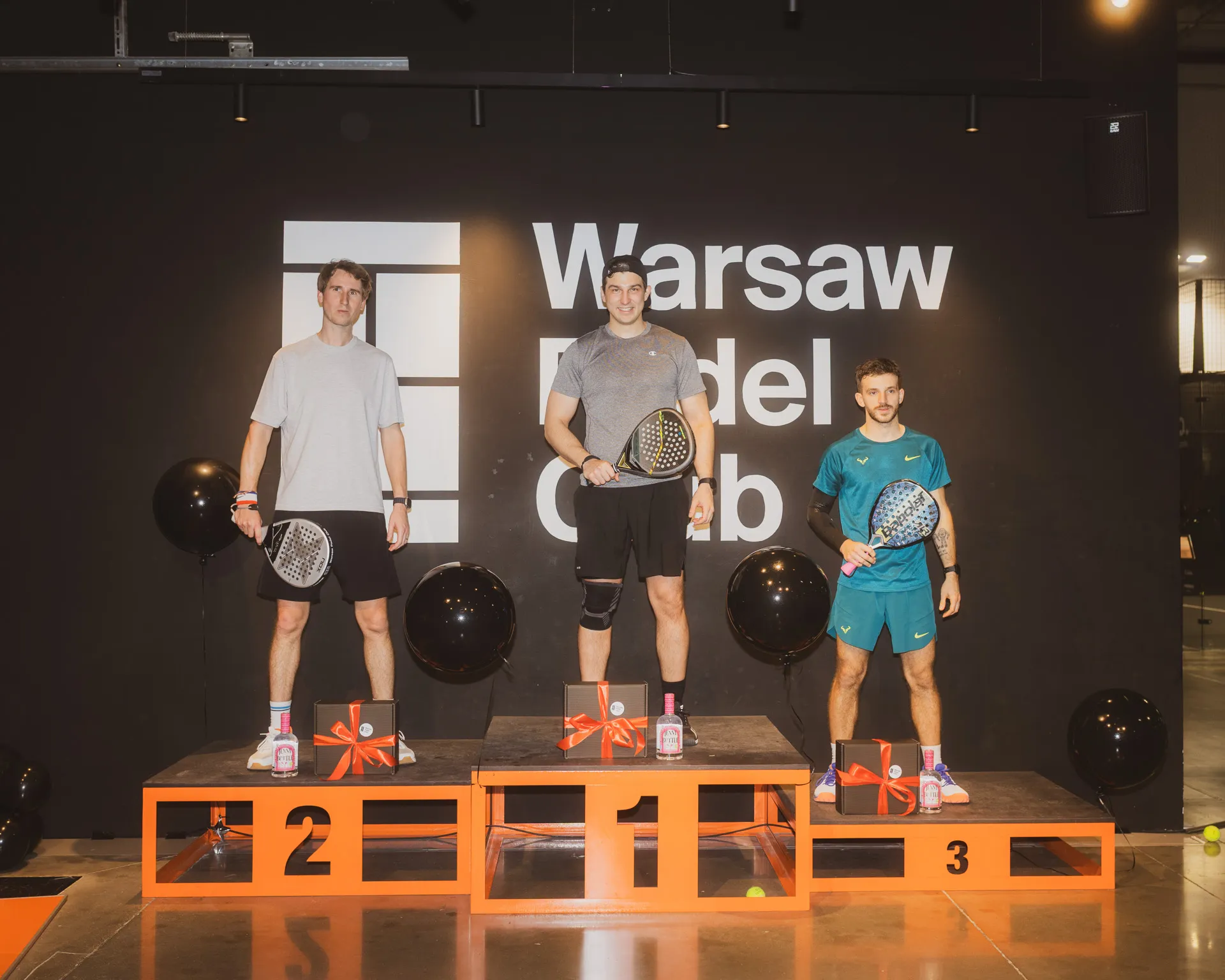 Three men holding padel rackets standing on winners podium in front of a Warsaw Padel Club sign with black balloons around.