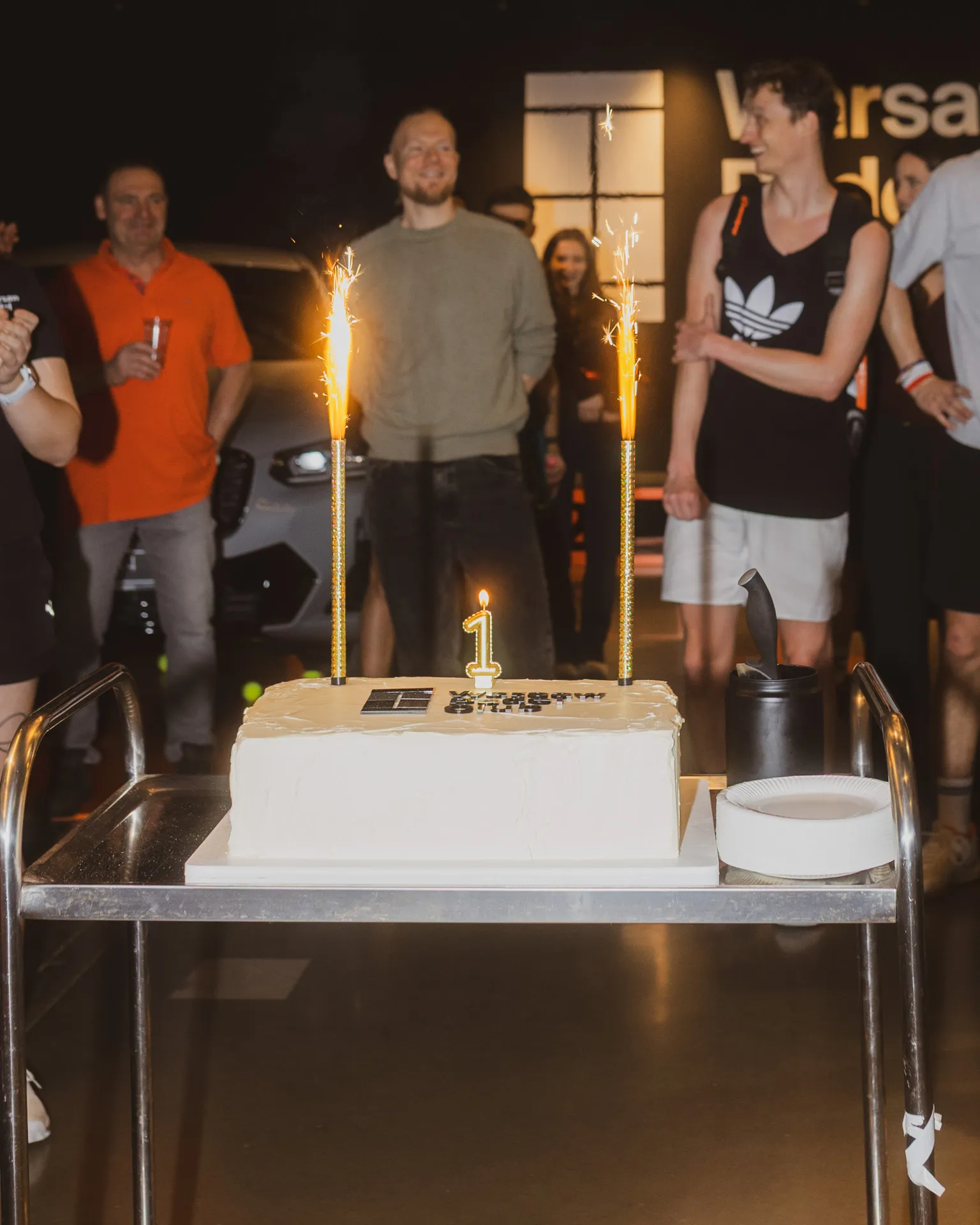 White rectangular cake with a '1' candle and two sparklers on a metal cart, people standing and smiling in the background.