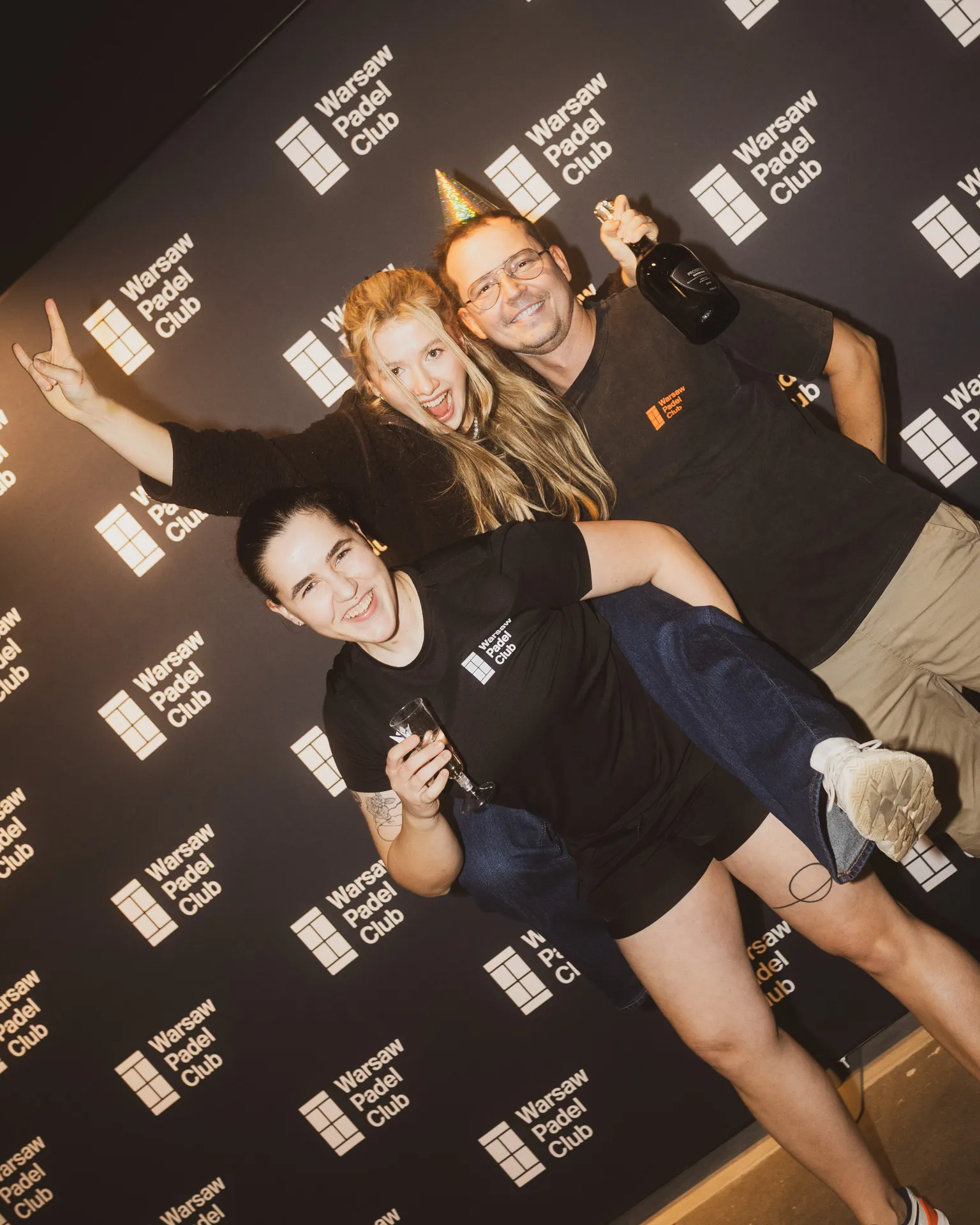 Three smiling people celebrating in front of a Warsaw Padel Club backdrop, one wearing a party hat and holding a bottle, another holding a glass.