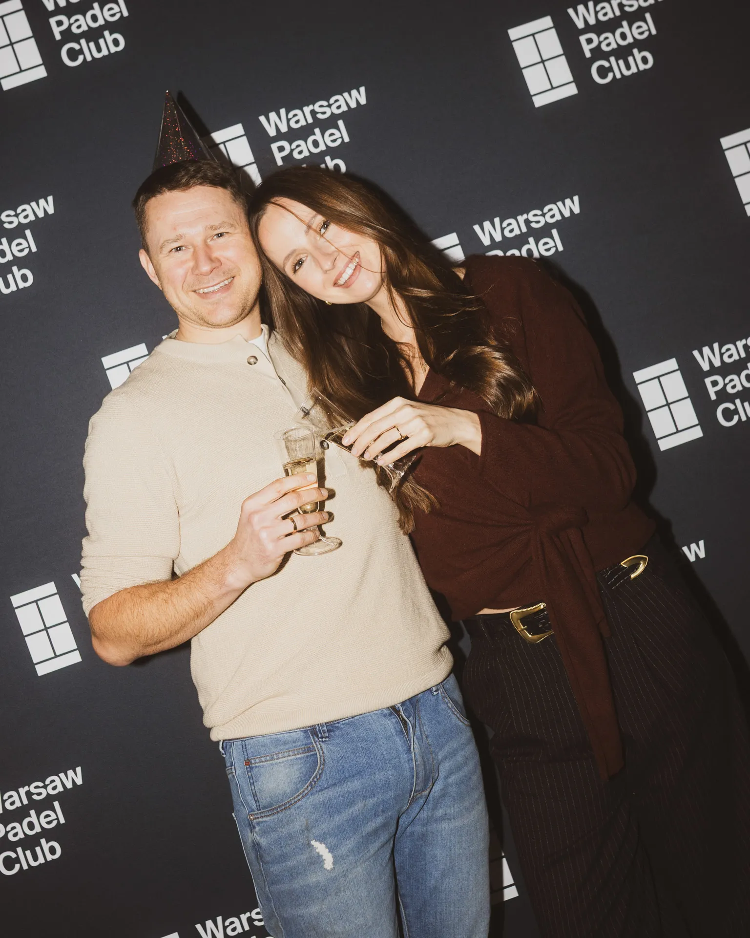 Smiling man wearing a party hat and a woman with long hair standing close together, each holding a glass of champagne, with a Warsaw Padel Club backdrop.