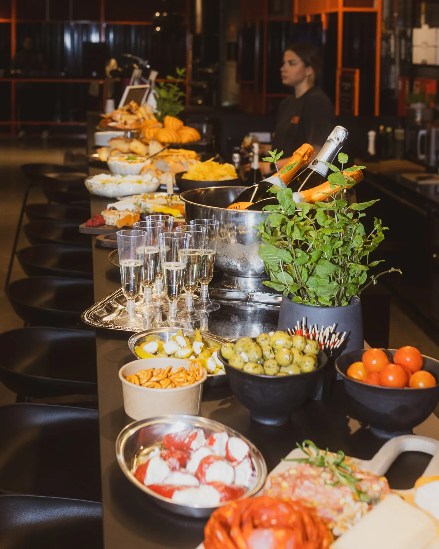 Buffet table with glasses of champagne, bottles in ice bucket, bowls of olives, cherry tomatoes, pretzels, and assorted appetizers with a woman in the background.