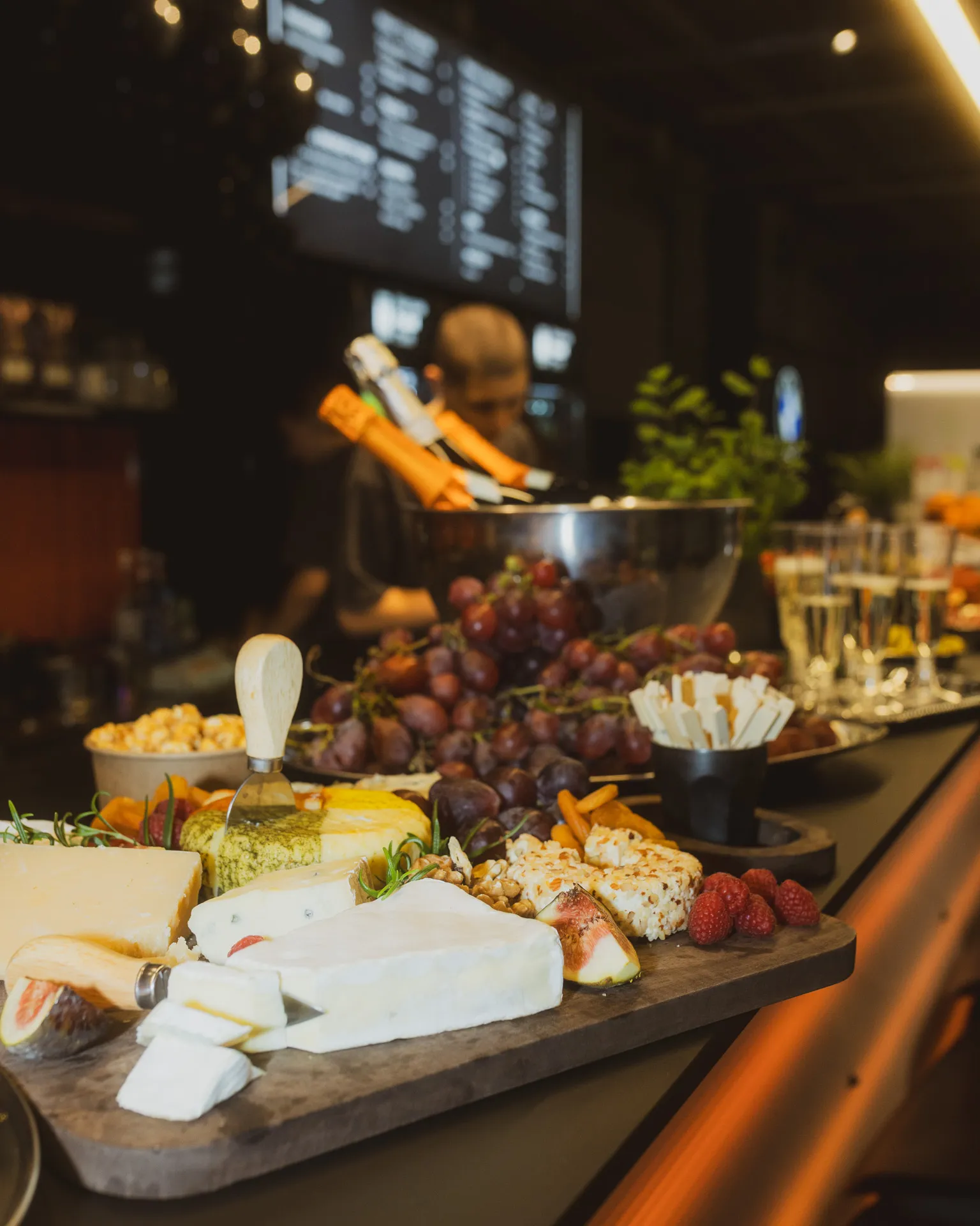 Close-up of a charcuterie board with assorted cheeses, fresh grapes, figs, raspberries, nuts, and crackers on a restaurant counter with blurred background.