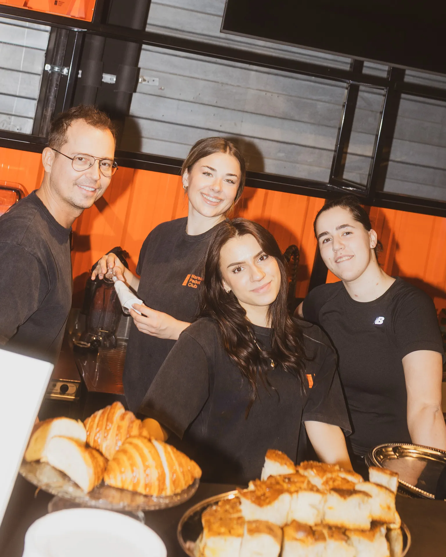 Four smiling people behind a table with croissants and bread, wearing black shirts, in a bright indoor setting.
