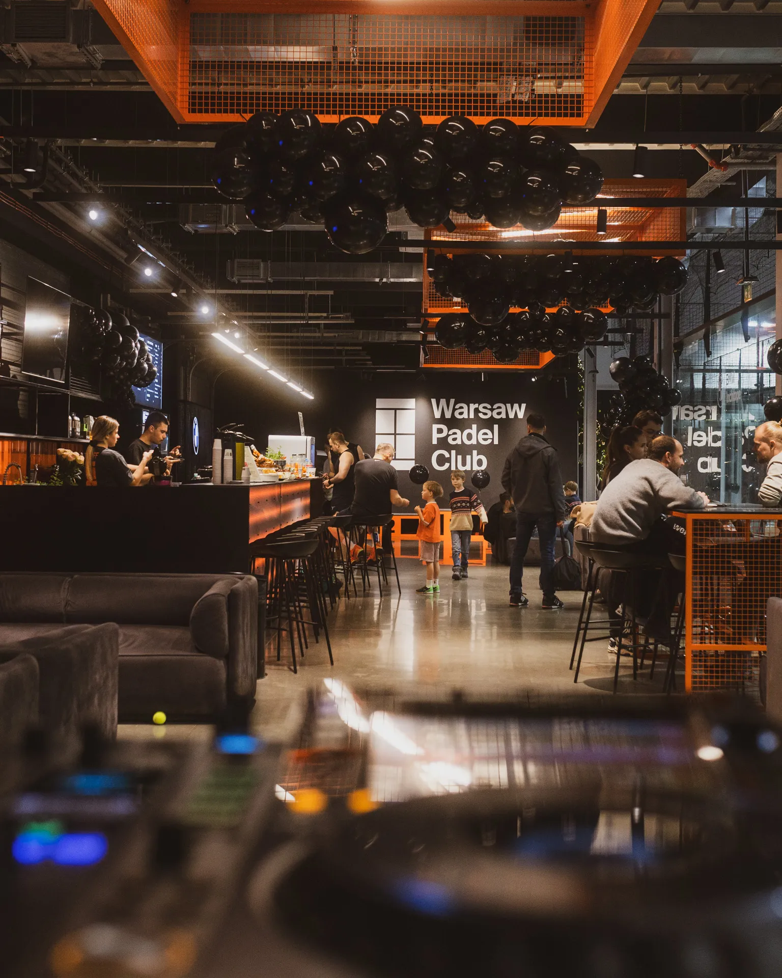 Interior of Warsaw Padel Club with people socializing near a bar, black balloons hanging from the ceiling, and children standing in the center.
