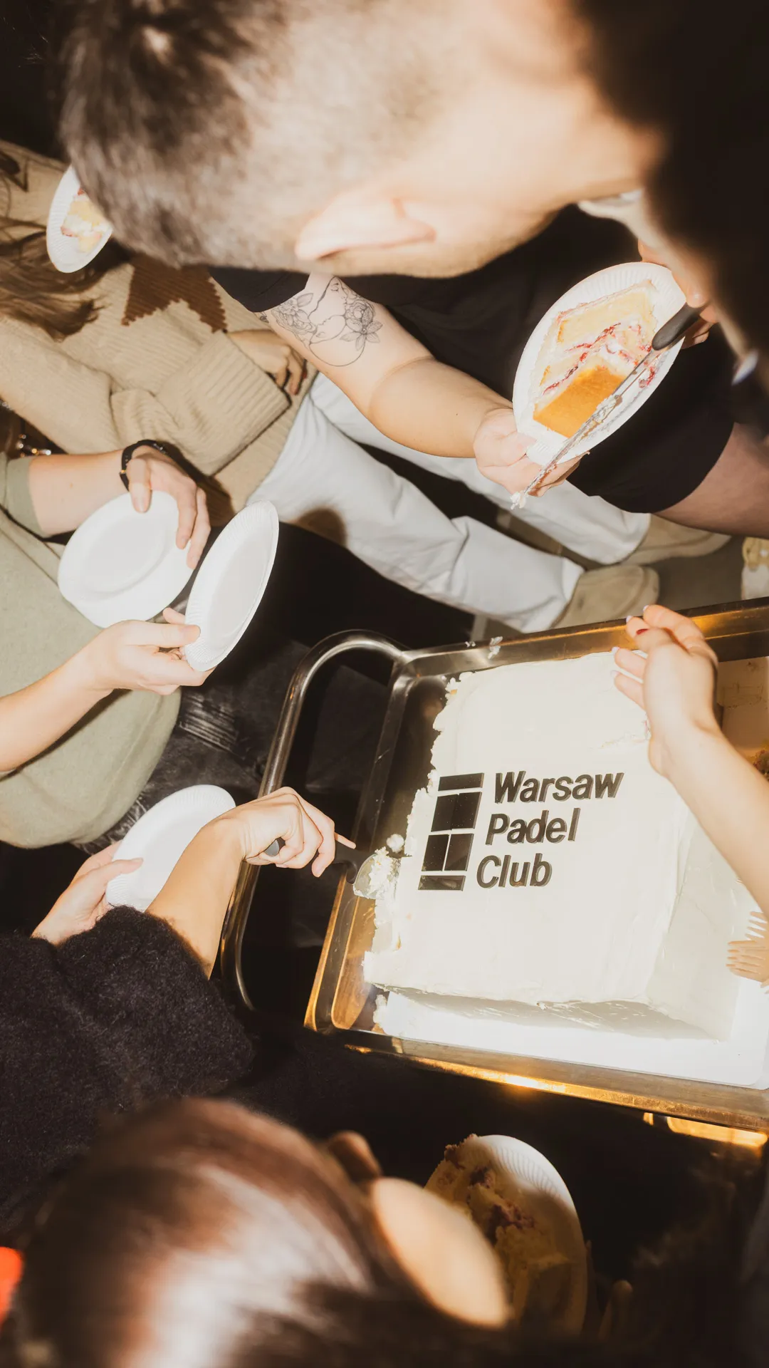 People serving and holding plates around a white rectangular cake with the Warsaw Padel Club logo.