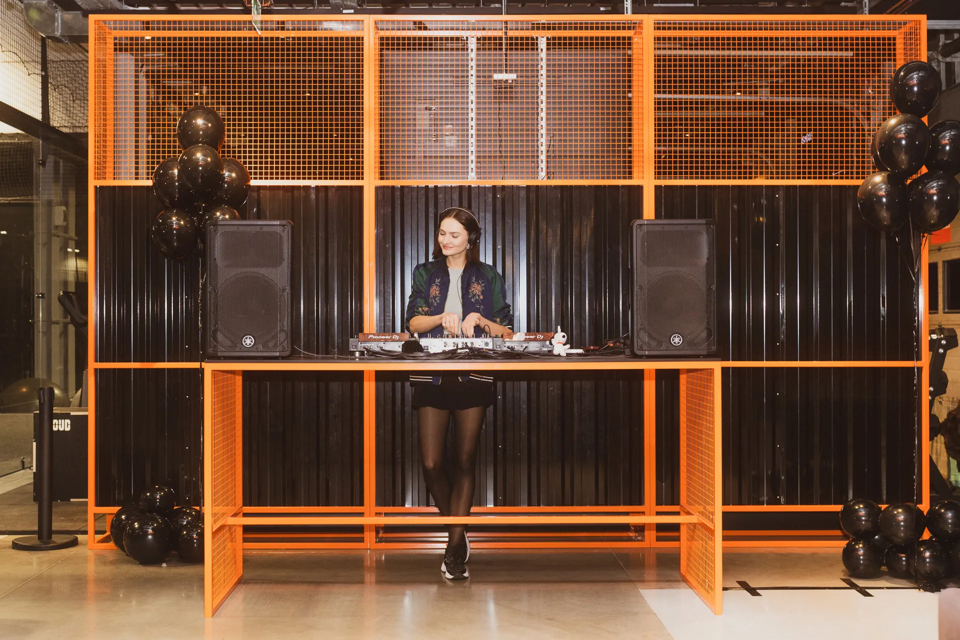 Woman DJ standing behind an orange metal table with mixing equipment, flanked by black speakers and black balloons.