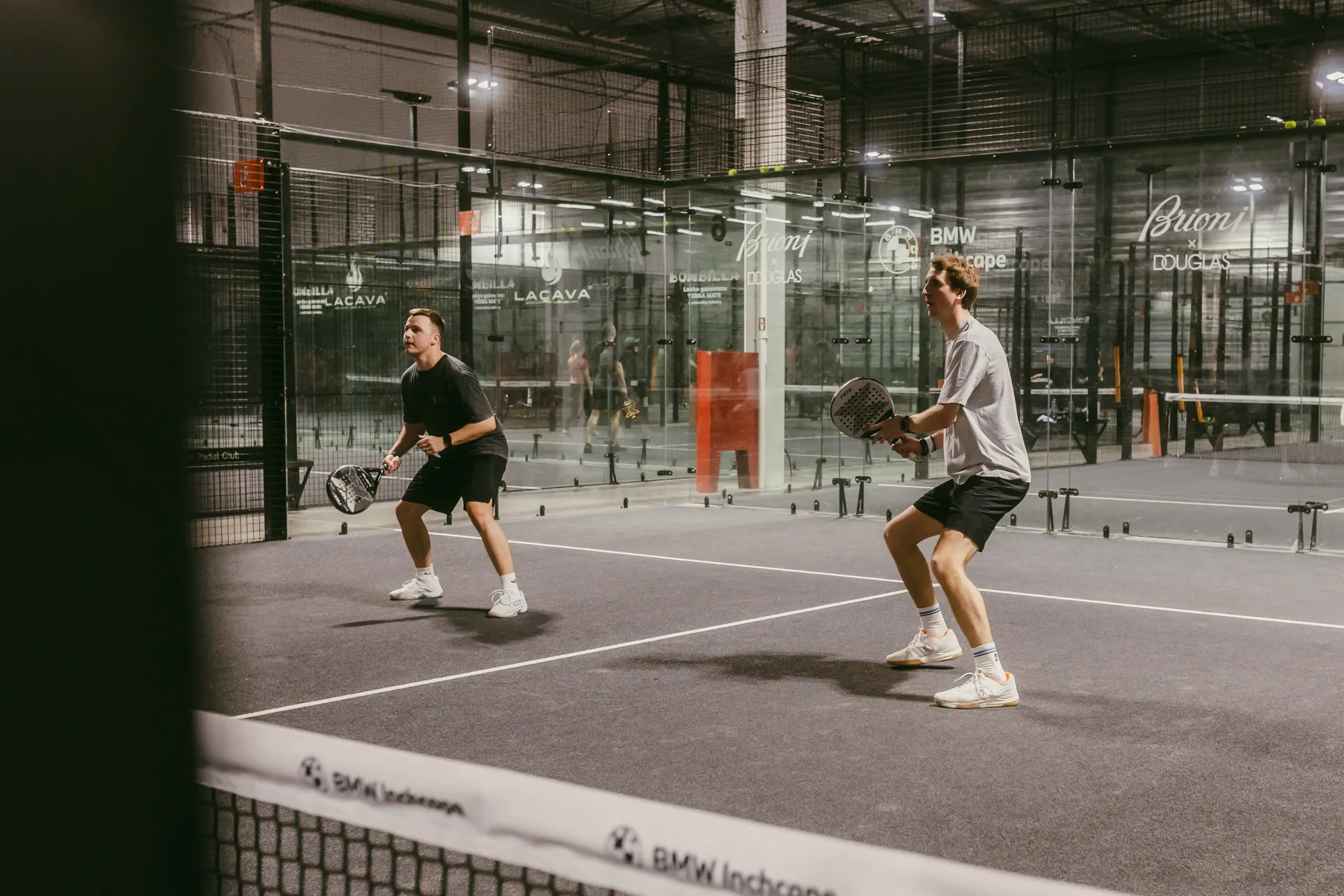 Two men playing padel on an indoor court, each holding a padel racket, preparing to return a shot.