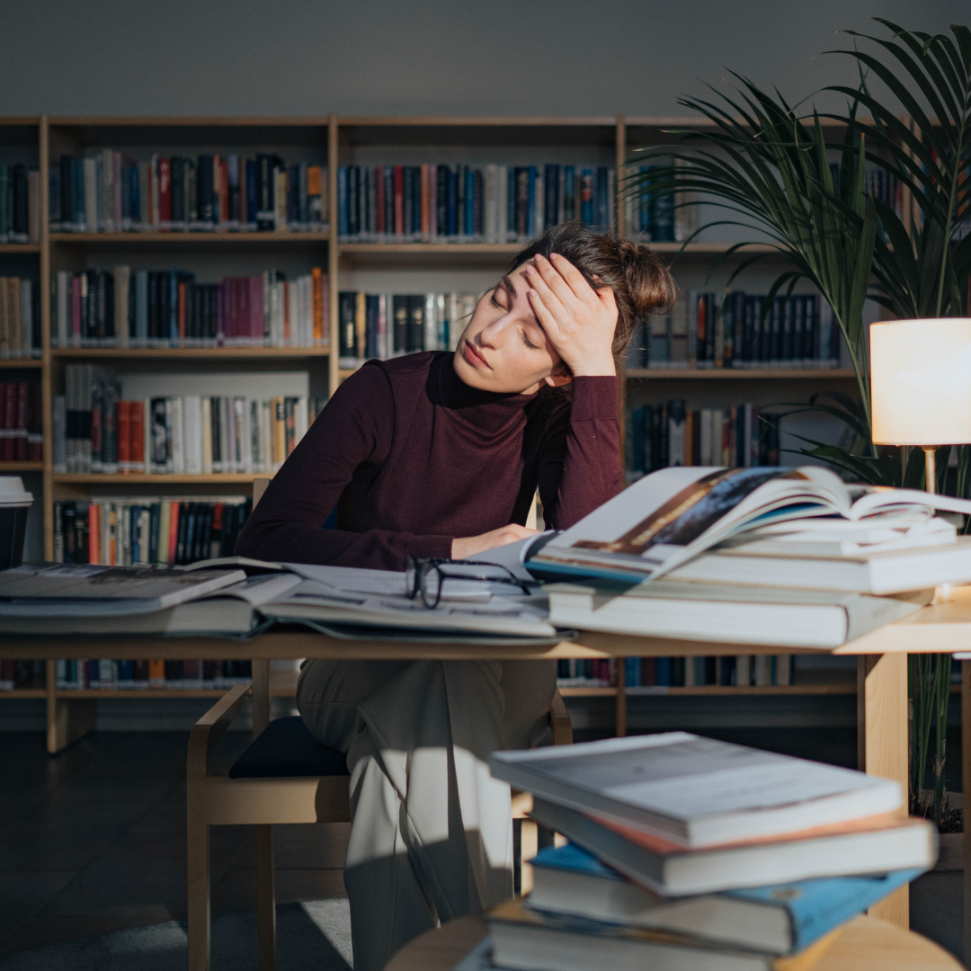 Tired woman in a purple sweater sitting at a table surrounded by open books in a library.