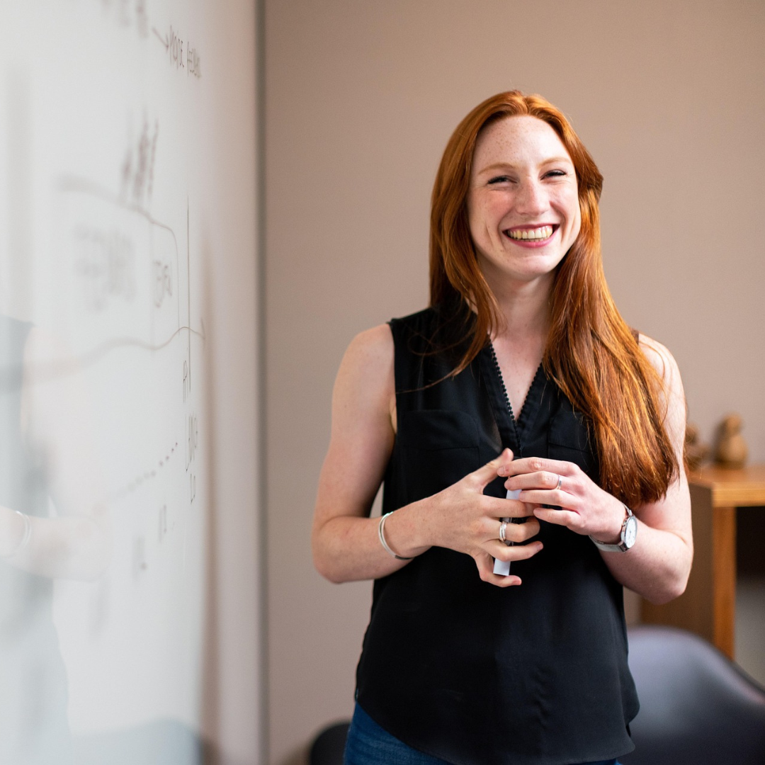 Smiling red-haired woman holding a marker standing next to a whiteboard with diagrams.