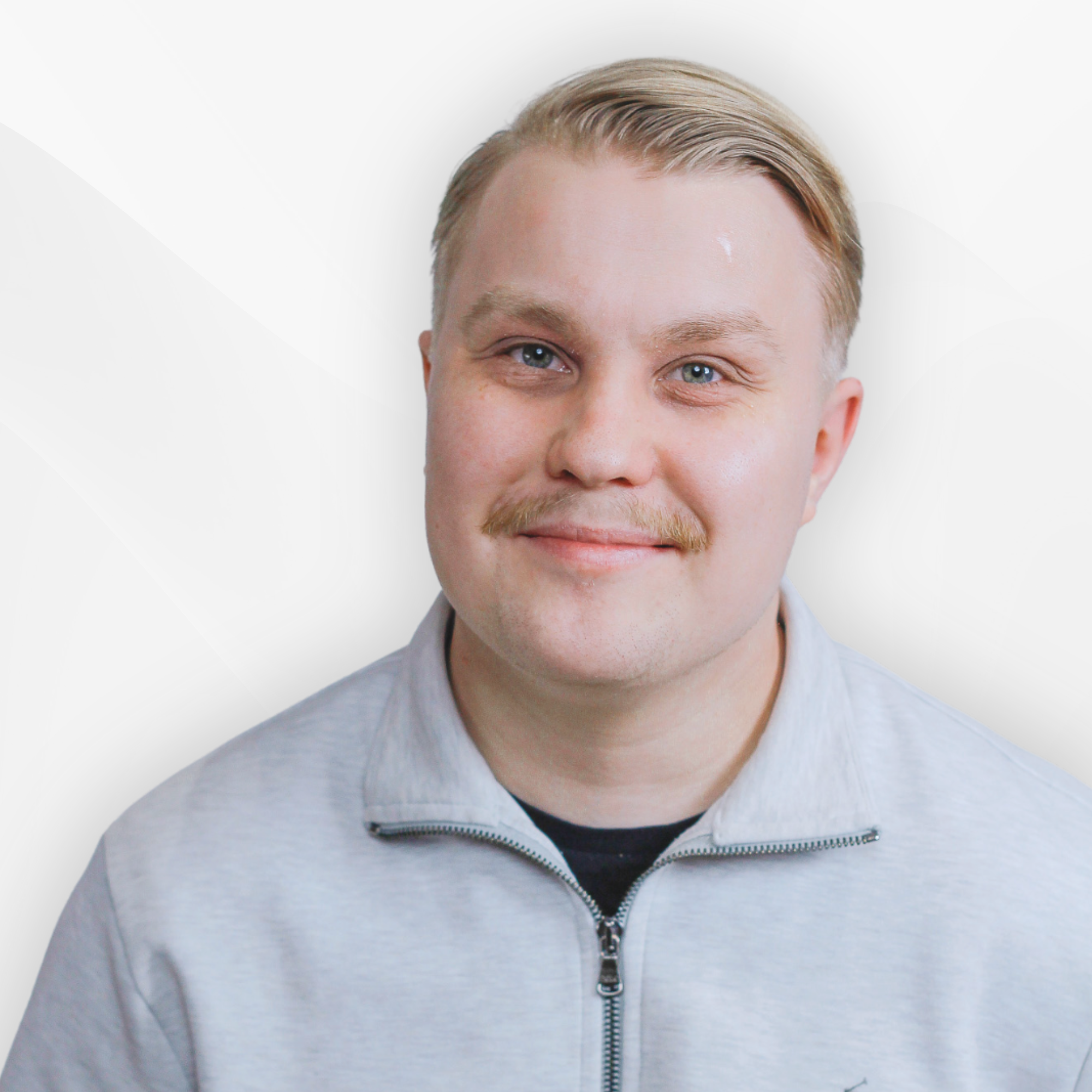 Smiling young man with blond hair and a mustache wearing a light gray zip-up jacket over a black shirt.