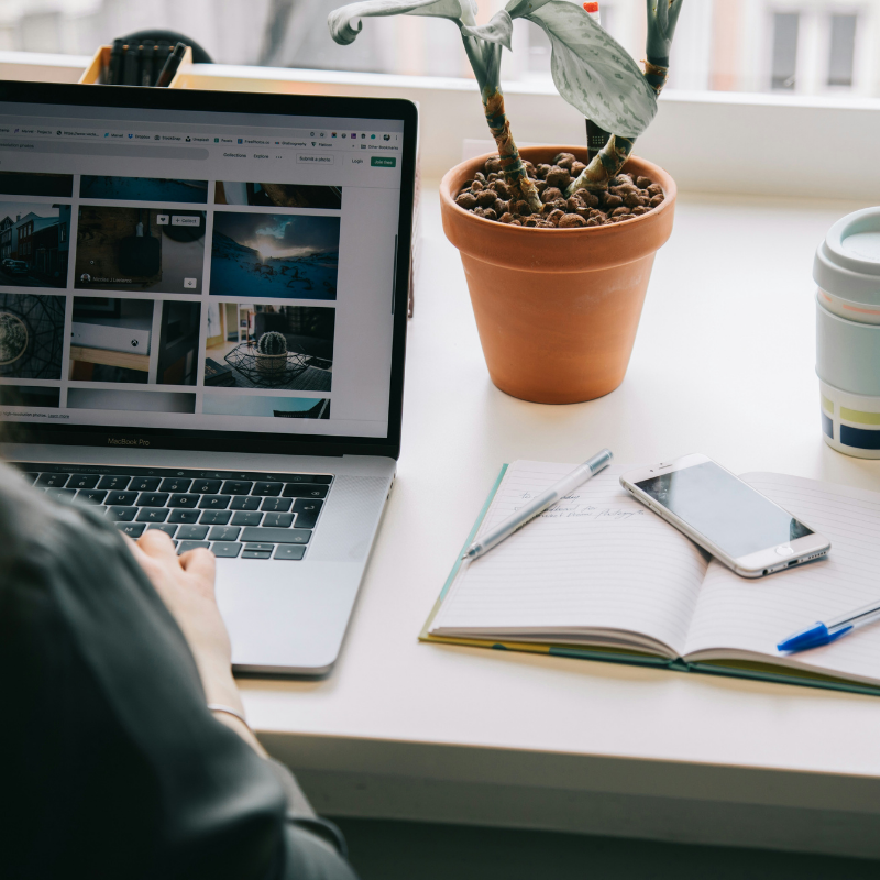 Person working on a laptop at a desk with an open notebook, smartphone, pen, potted plant, and reusable coffee cup near a window.