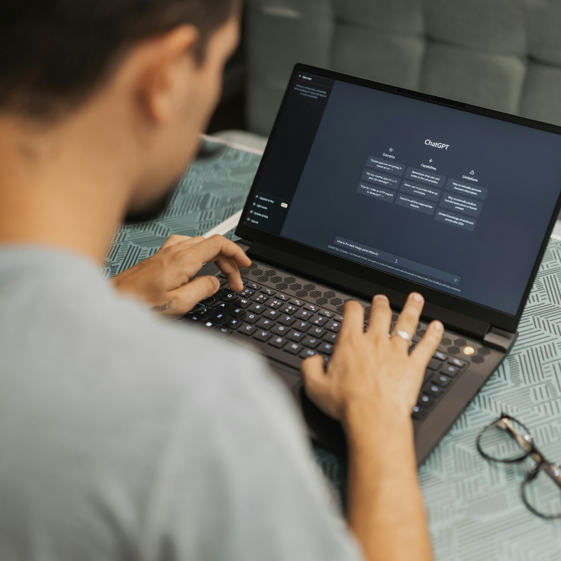 Person typing on a laptop displaying the ChatGPT interface with a patterned tablecloth and glasses nearby.