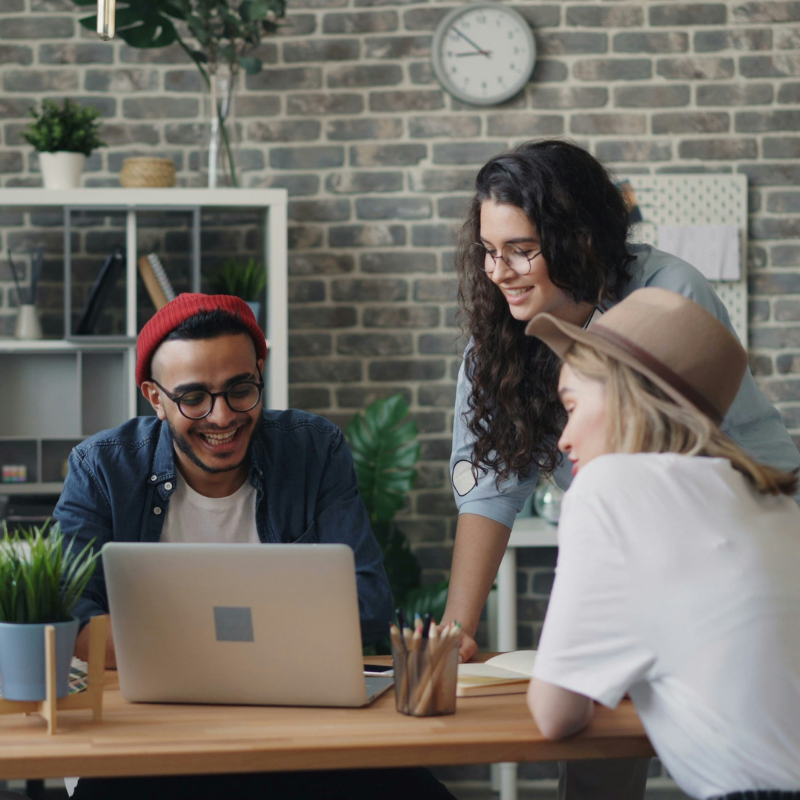 Three young people smiling and collaborating while looking at a laptop on a wooden desk in a modern office.