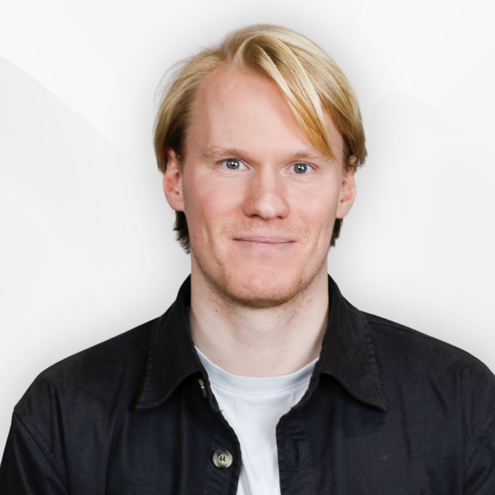 Young man with blonde hair wearing a black jacket over a white shirt, smiling slightly against a plain light background.