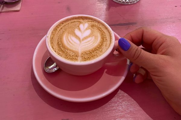 Hand with purple nail polish holding a cup of latte with leaf latte art on a pink saucer and table.