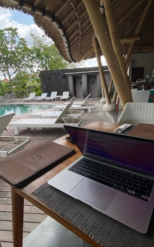 Laptop on a table by a pool with lounge chairs under a thatched roof structure.