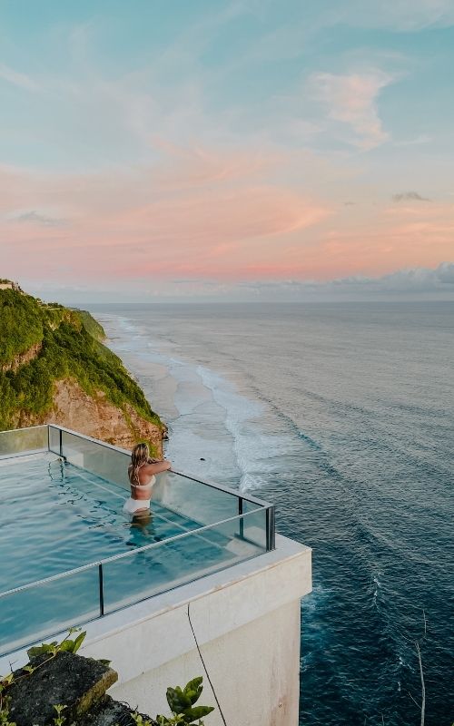 Woman in a rooftop pool overlooking a coastal cliff and calm ocean at sunset.