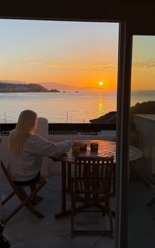 Person sitting on a balcony at sunset with a sea and mountain view, two drinks on wooden table.