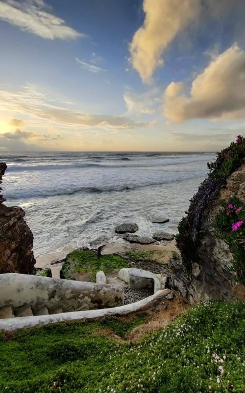 Stone stairway leads down to rocky beach with ocean waves under a cloudy sky at sunset.