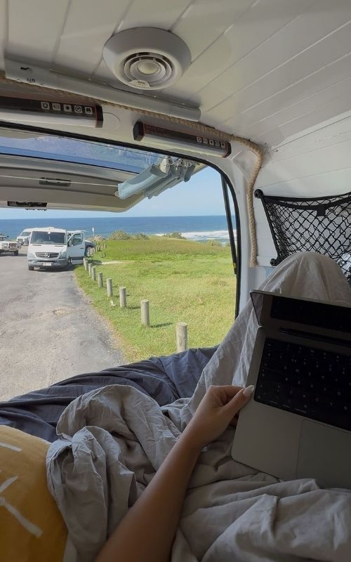 Person inside a van using a laptop, with an open door showing grass, cars, and ocean in the distance.