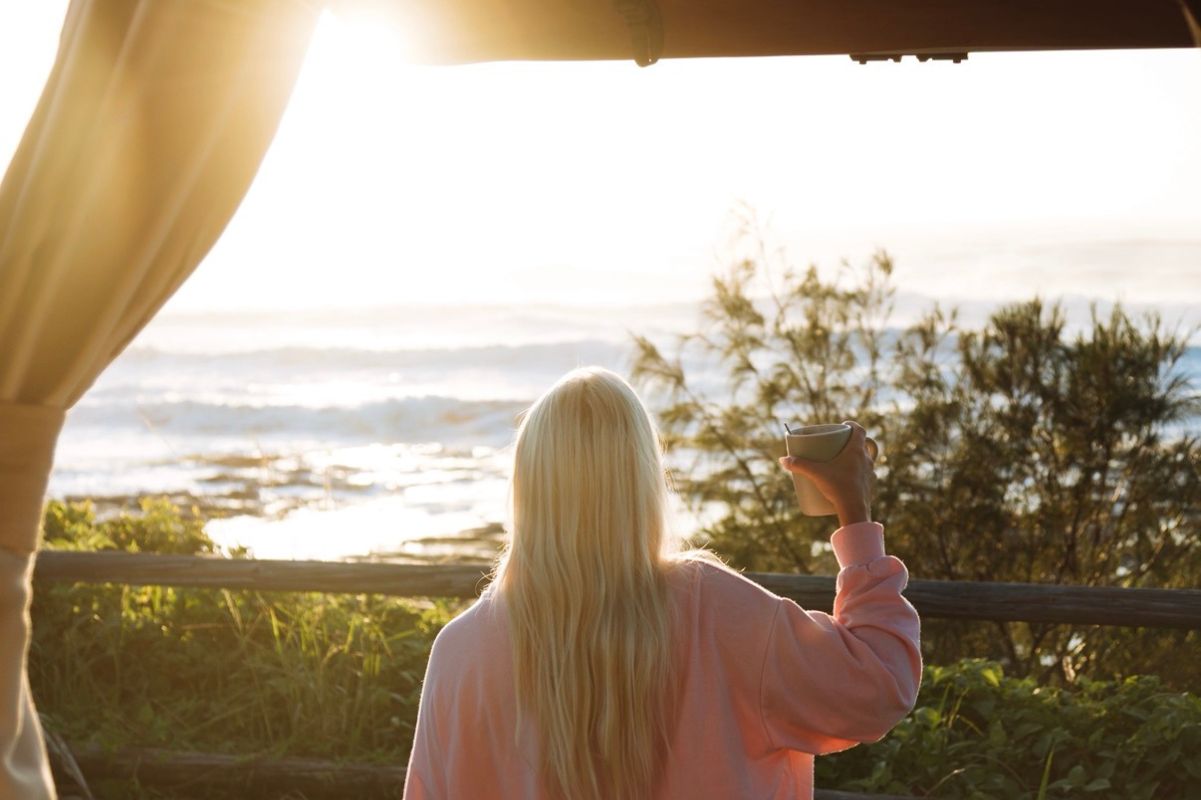 Person with long blonde hair in pink sweater holding a cup overlooking sunlit ocean and greenery.