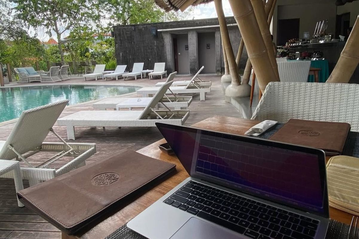 Outdoor workspace by a pool with a laptop on a wooden table and white lounge chairs around the pool.