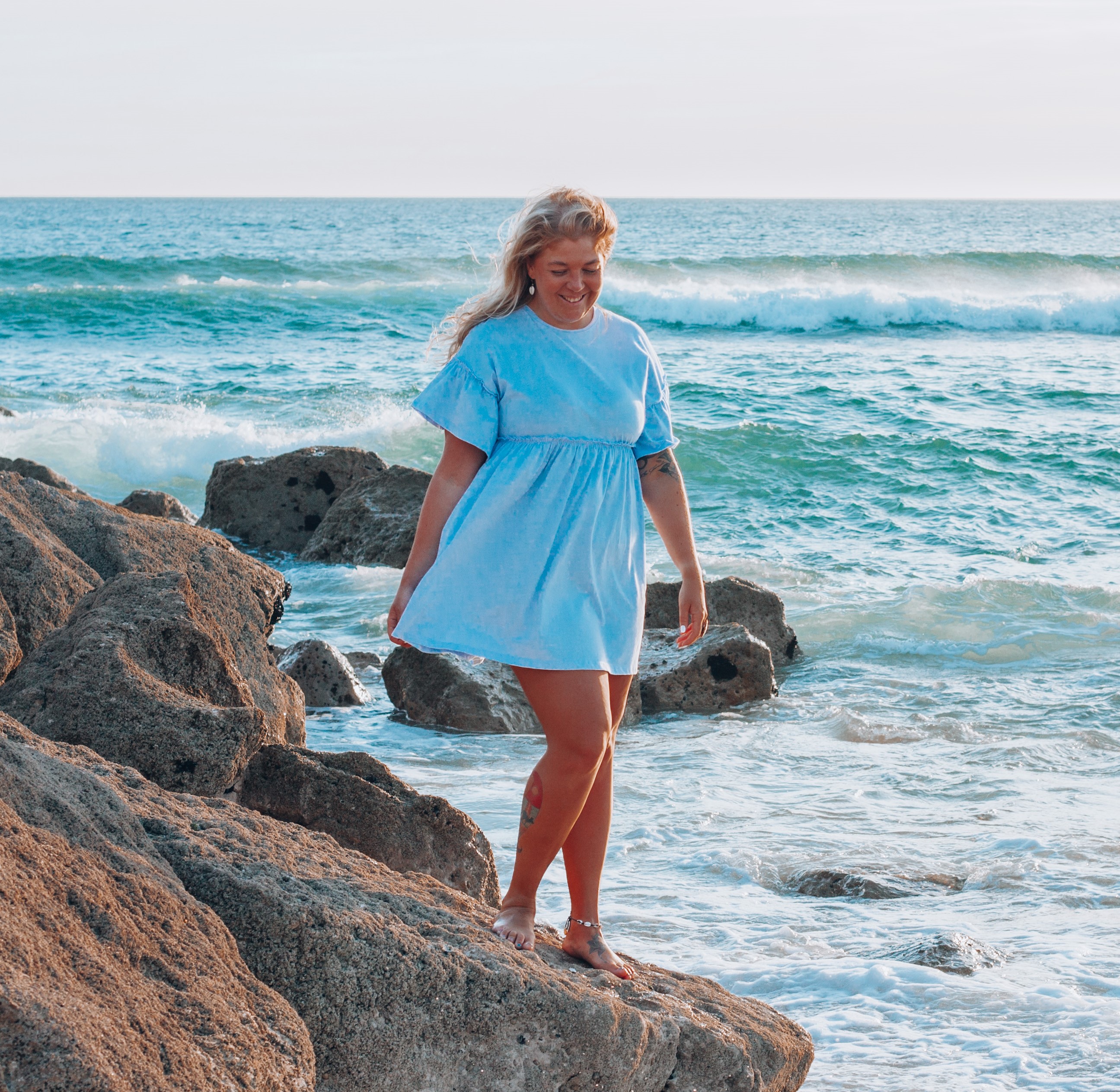 Smiling woman in a blue dress walking barefoot on rocks by the ocean waves at sunset.