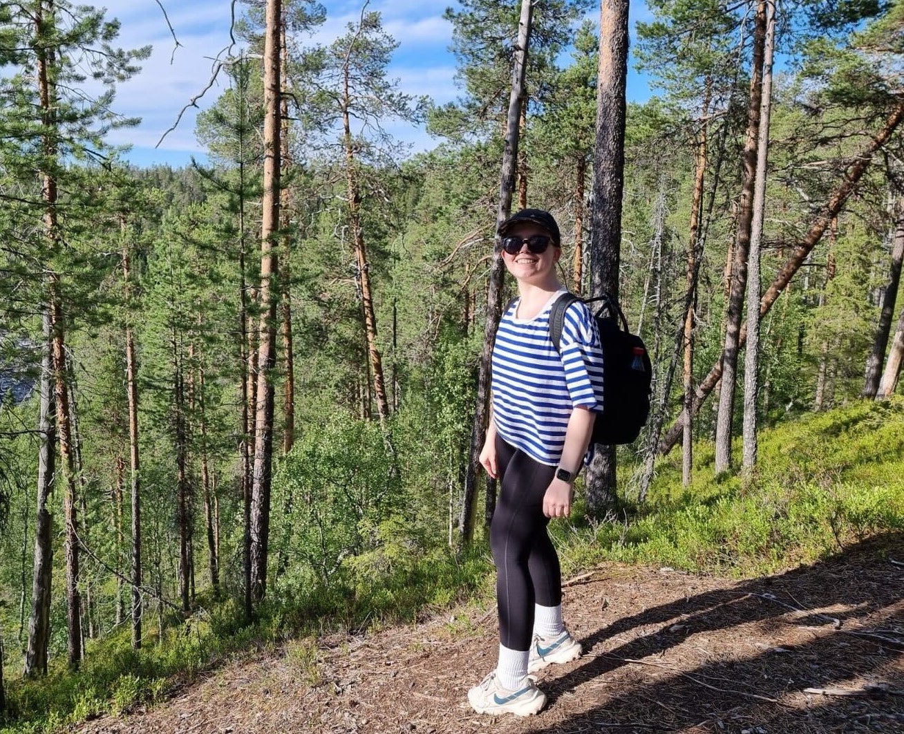 Smiling woman with backpack and sunglasses standing on forest trail among pine trees in sunny weather.