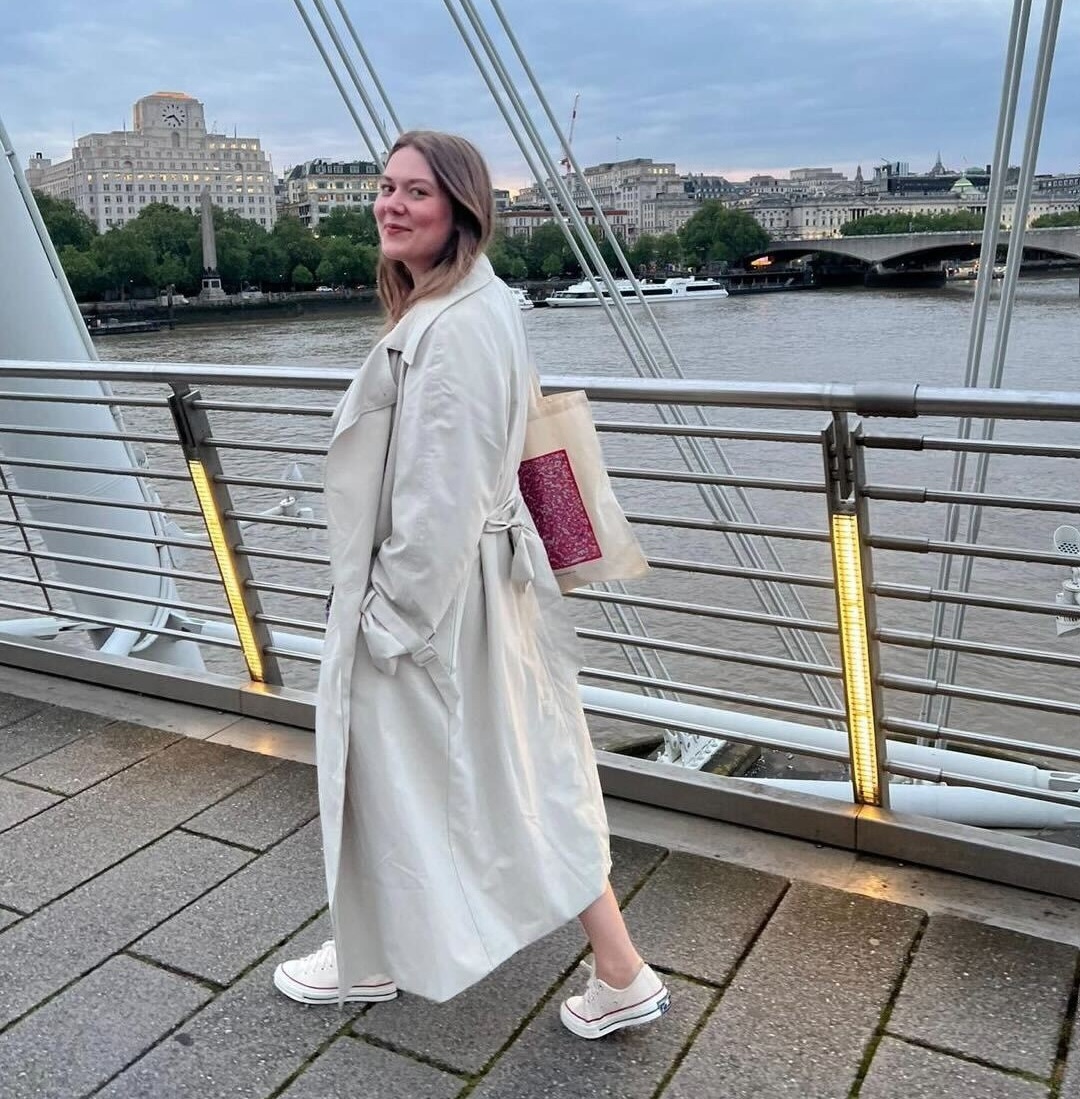 Woman in a light trench coat and white sneakers standing on a bridge over a river at dusk.