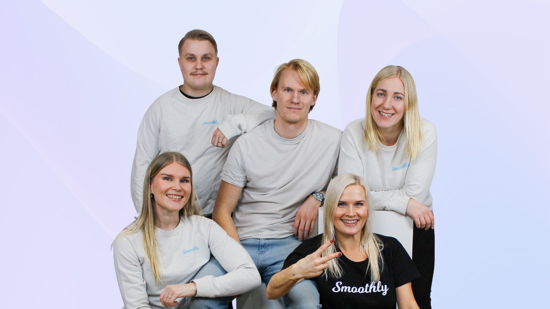 Group of five smiling young adults wearing Smoothly branded shirts posing against a gray background.