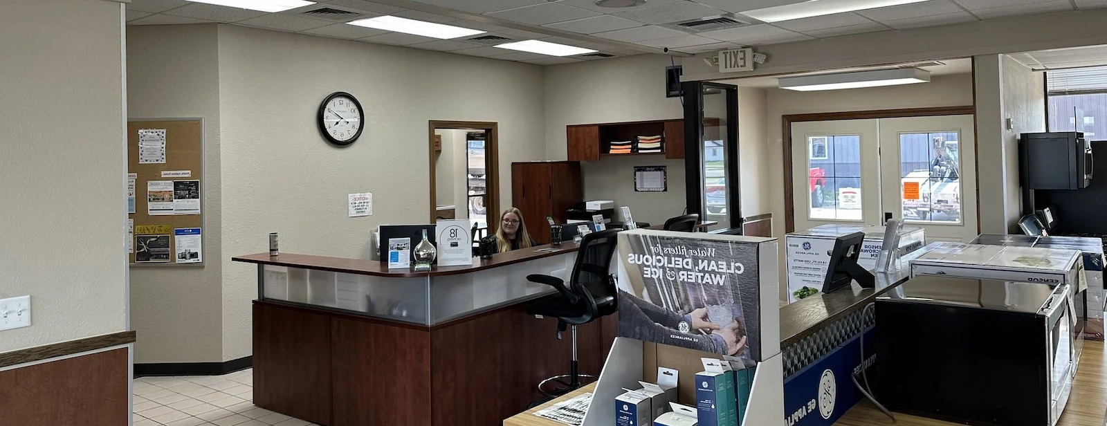 Reception area with a woman sitting behind a curved wooden desk, bulletin board, clock on the wall, and office equipment near the entrance.