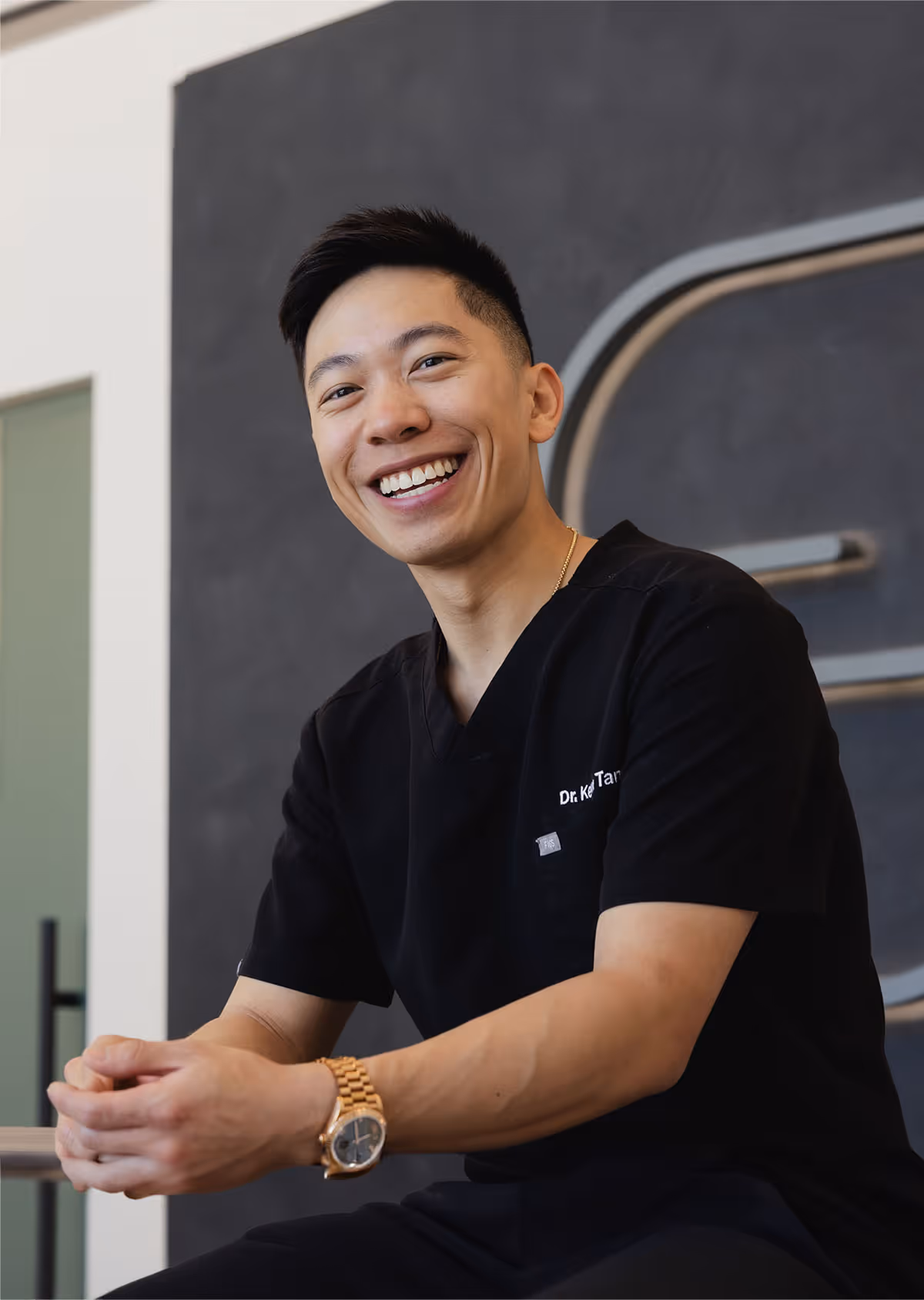 Smiling man in black scrubs with rolled-up sleeves and gold watch sitting indoors.
