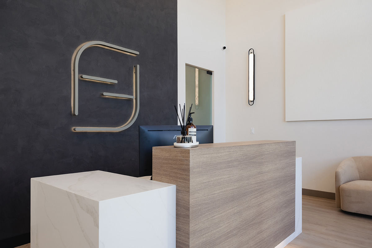 Modern reception desk with wood and white marble finish, black wall with logo, and beige chair nearby.