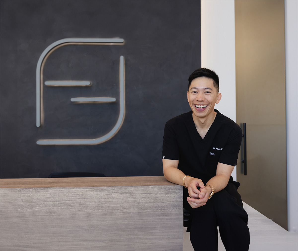 Smiling man in black scrubs leaning on a light wooden counter with a dark wall and logo behind him.