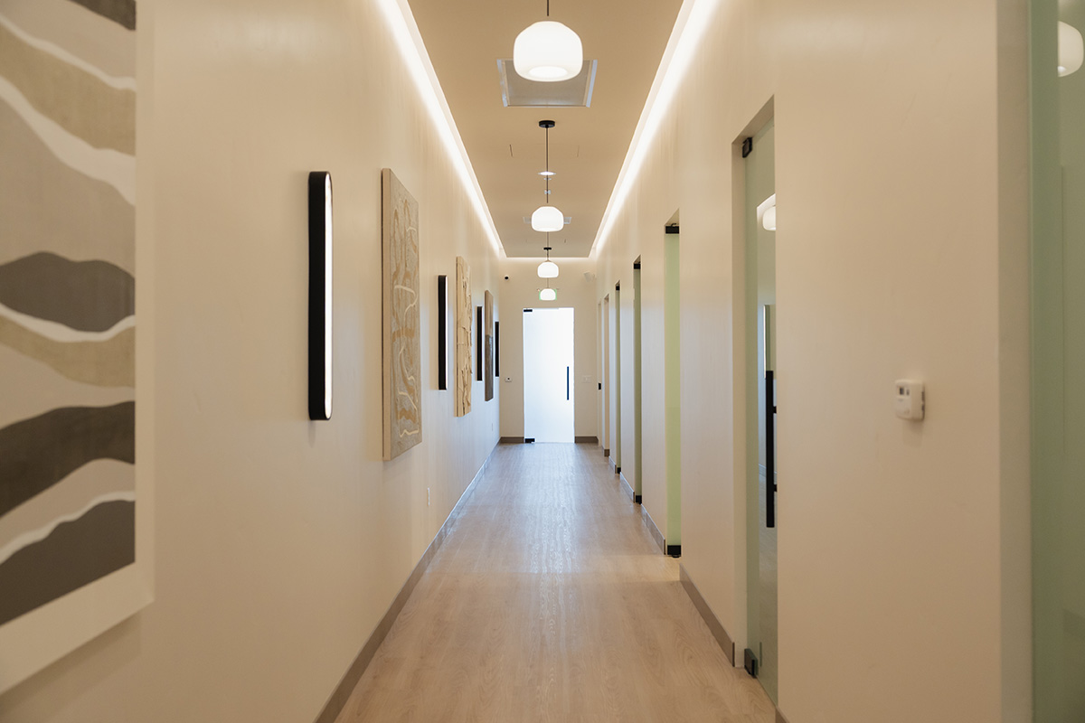 Long, well-lit beige hallway with framed abstract art on one side and glass doors on the other.