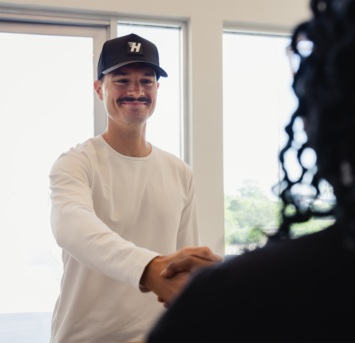 Man with mustache wearing a black cap and white shirt shaking hands with another person indoors.