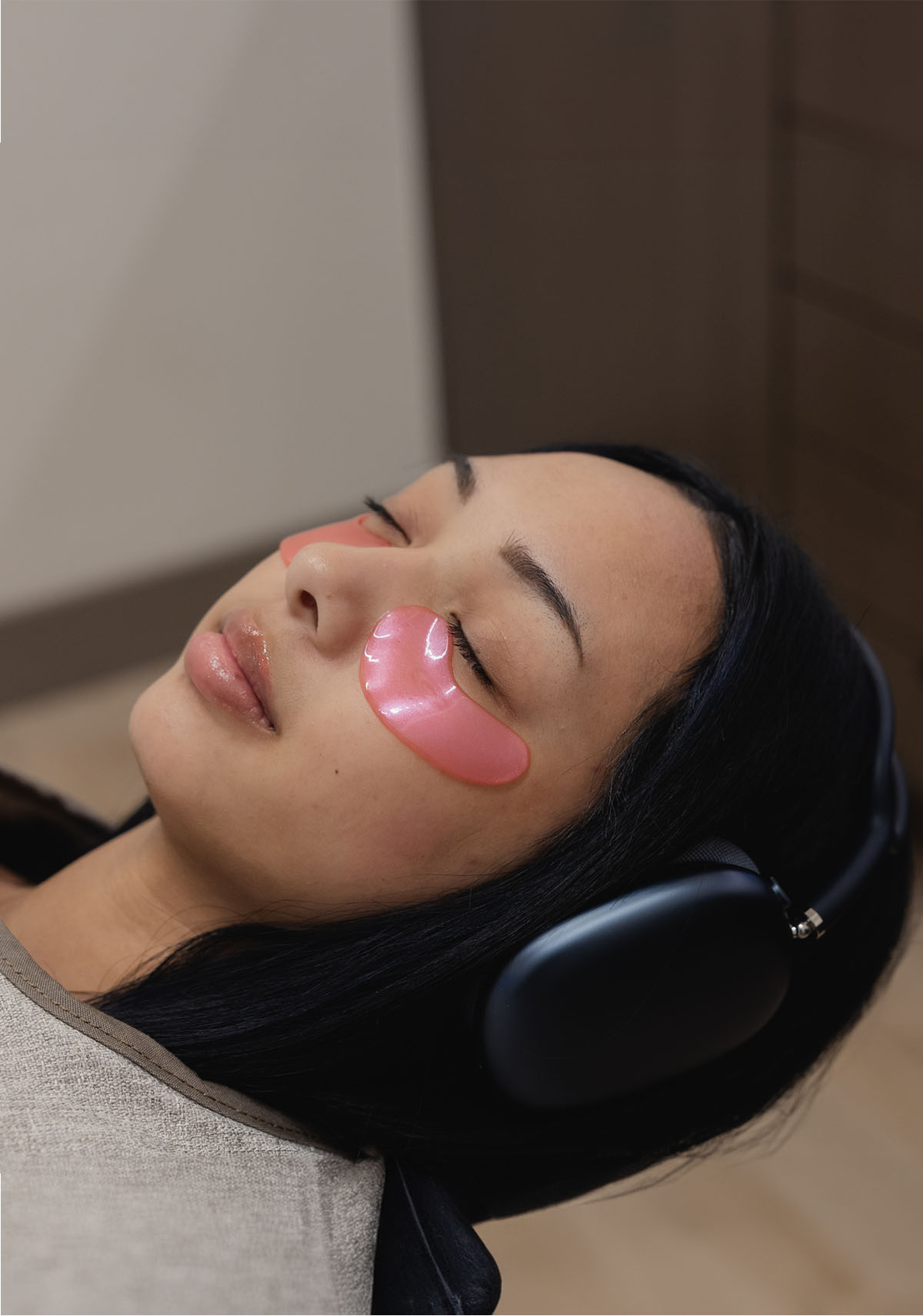 Young woman reclined with pink under-eye gel patches and black over-ear headphones, eyes closed.