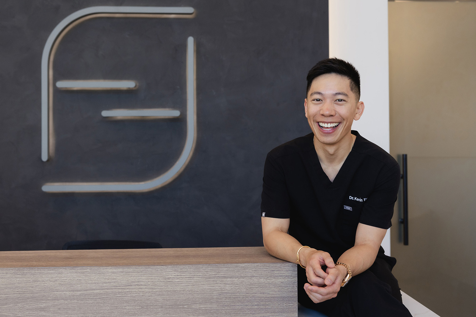 Smiling male doctor in black scrubs sitting at a wooden reception desk with a modern logo on wall.
