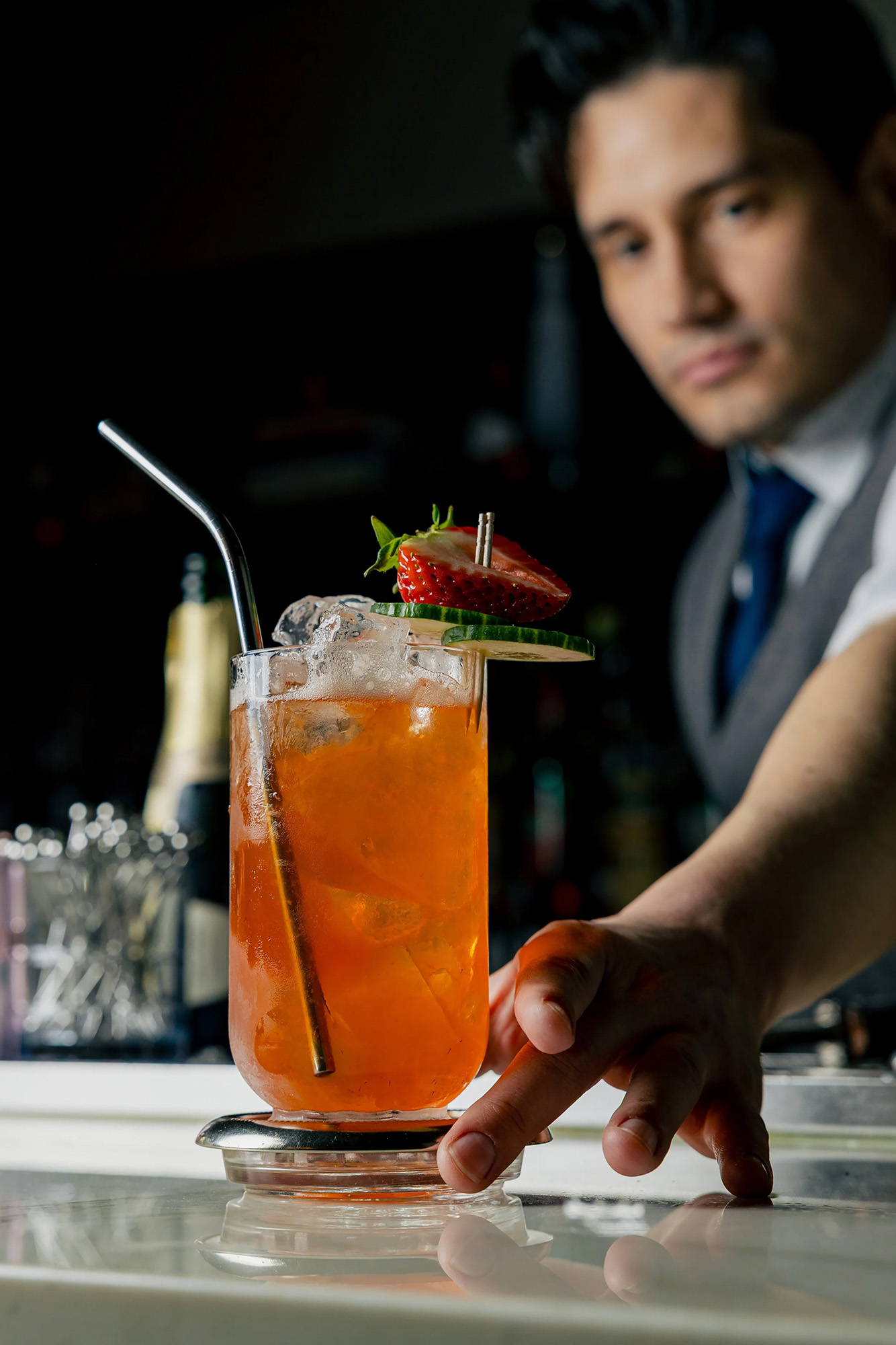 Bartender serving an iced orange cocktail garnished with strawberry and cucumber slice with a metal straw.