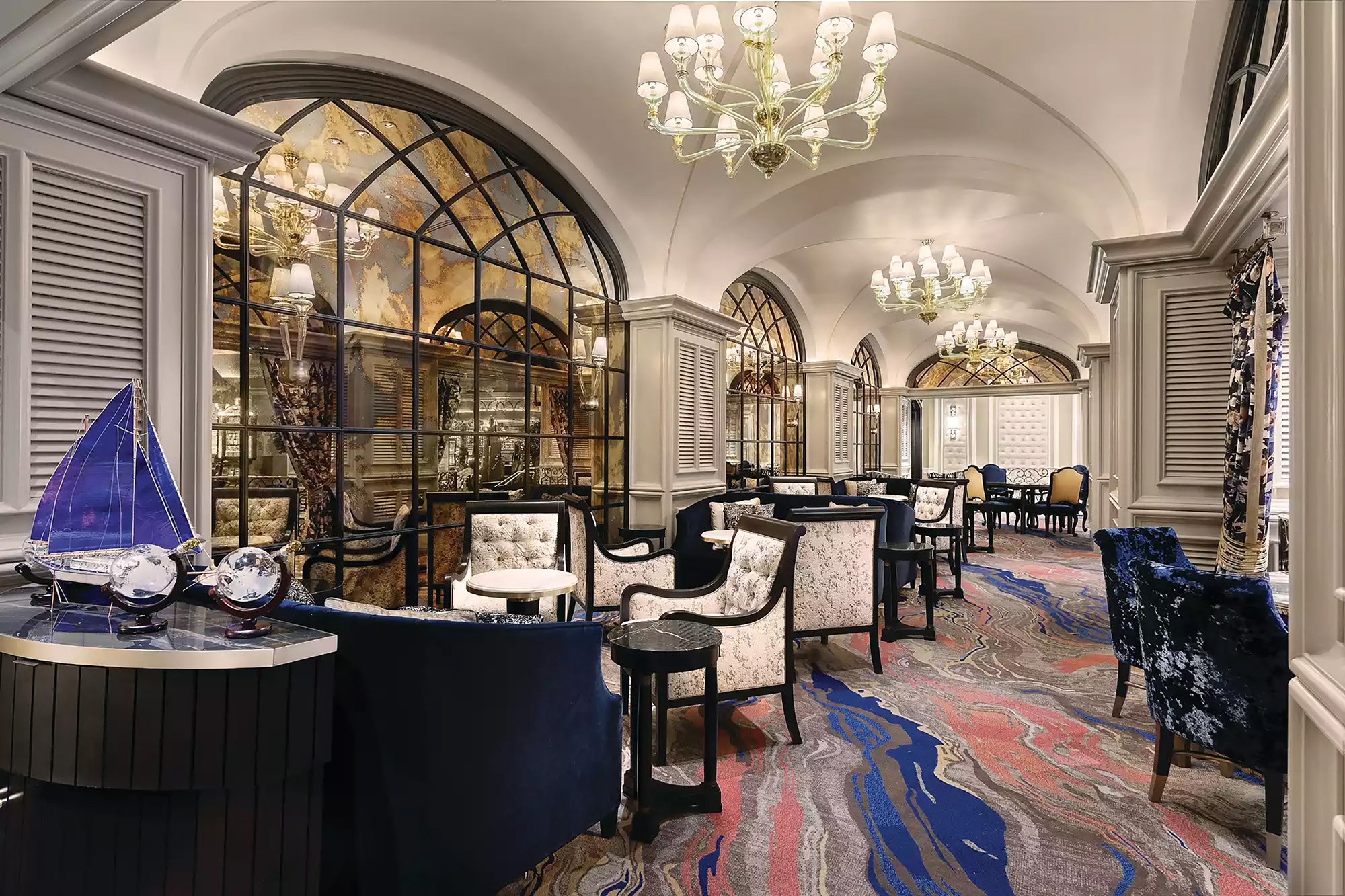 Elegant lounge with arched mirrors, chandeliers, patterned carpet, blue and floral upholstered chairs.