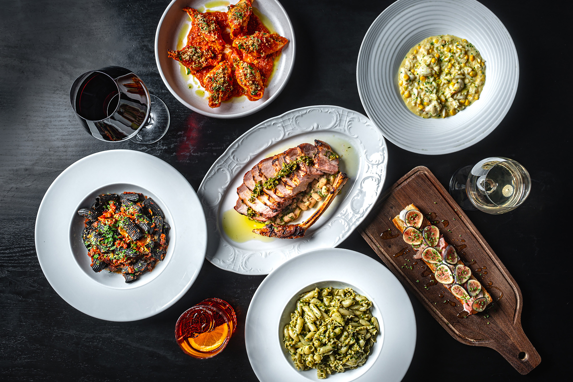 Top view of assorted gourmet dishes with glasses of red, white wine, and a cocktail on black table.