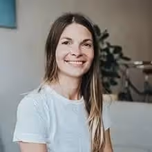 Frau mit langem braunem Haar und weißem T-Shirt lächelt in einem hellen Raum mit Pflanze im Hintergrund.