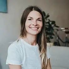 Frau mit langem braunem Haar und weißem T-Shirt lächelt in einem hellen Raum mit Pflanze im Hintergrund.